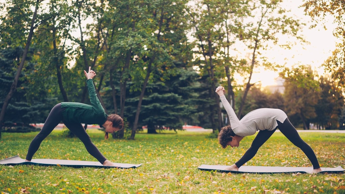 Female yoga student is exercising outdoors with instructor stretching legs and back bending forward standing on mats on green and yellow meadow in city park.