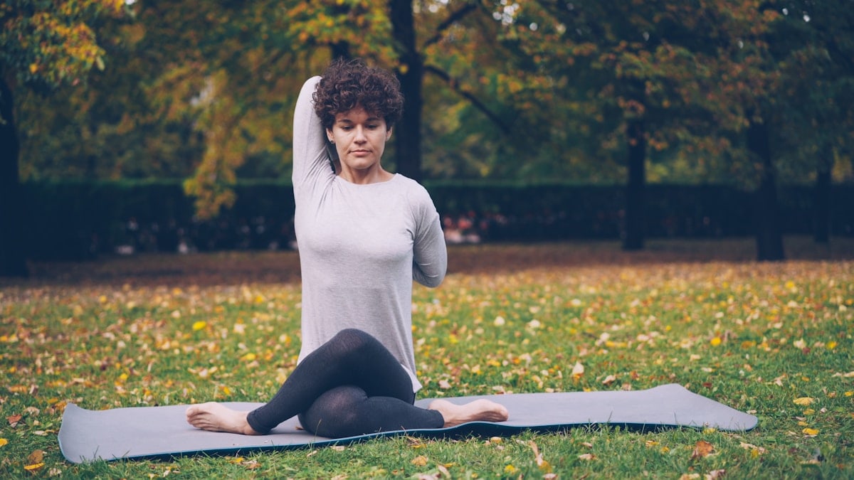 Serious young woman yogini is training in park sitting in Cow Face pose with arms behind her back during open-air practice in city park. Nature, youth and health concept.